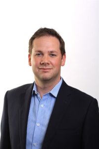 A professional headshot of Jake Morris wearing a dark suit jacket and blue striped shirt against a white background